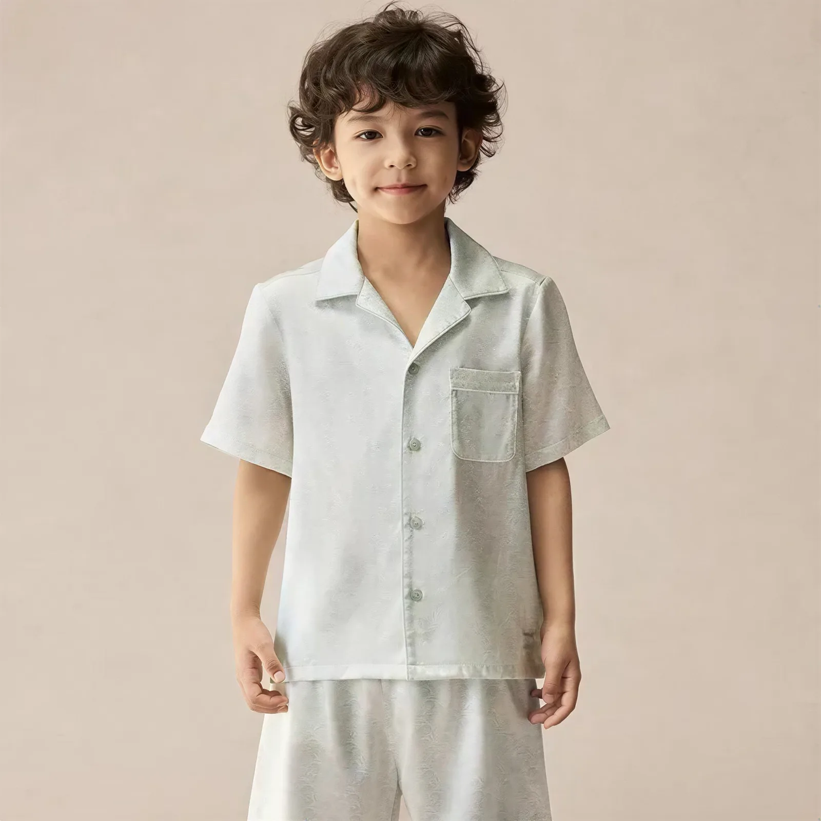 Kids private label silk pajama set detail showing a short-sleeve button-front shirt with notched collar, chest pocket, piped edge finish, straight hem